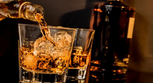 Whiskey in zwei Gläsern mit Eiswürfel fuellen mit Flasche im Hintergrund